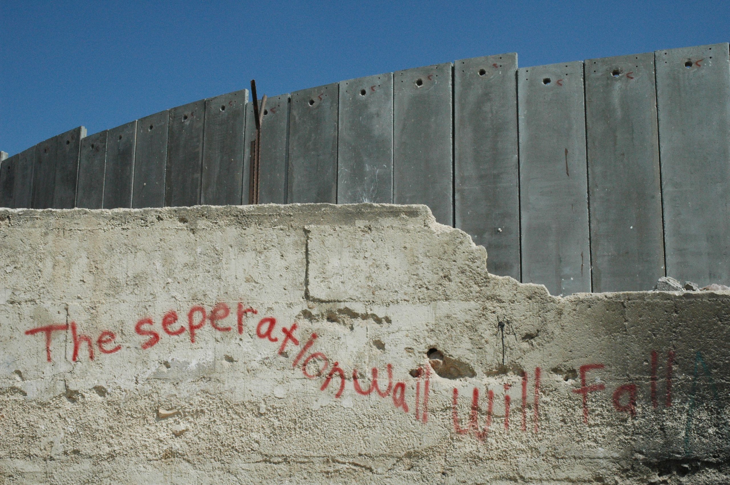 Photo taken in Palestine, showing the apartheid wall that divides the occupied lands. Two walls, one larger standing behind the other, that is breaking. There is graffiti on the front wall reading "the separation wall will fall"